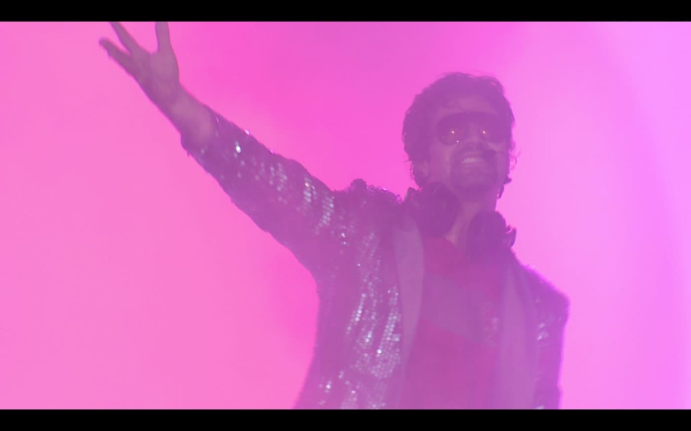 Daniel em palco, de casaco de lentejoulas e óculos escuros, com o braço no ar sob luz rosa.