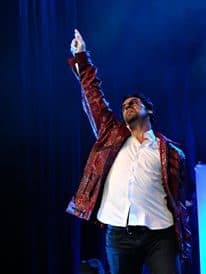 Daniel em palco, de casaco vermelho brilhante e braço levantado, sob luz azul.