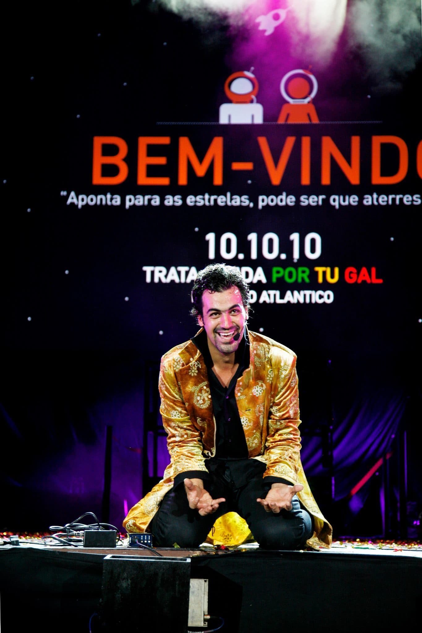 Daniel de joelhos em palco, de casaco dourado, à frente do painel "Bem-vindo — 10.10.10 — Trata a Vida Por Tu — Pavilhão Atlântico".