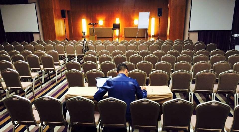 Sala de formação vazia antes do evento, com Daniel sentado ao portátil na última fila.